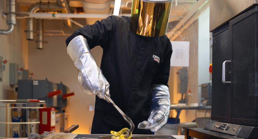 Man in lab wearing a safety mask and metallic gloves manipulates molten glass on a metal table